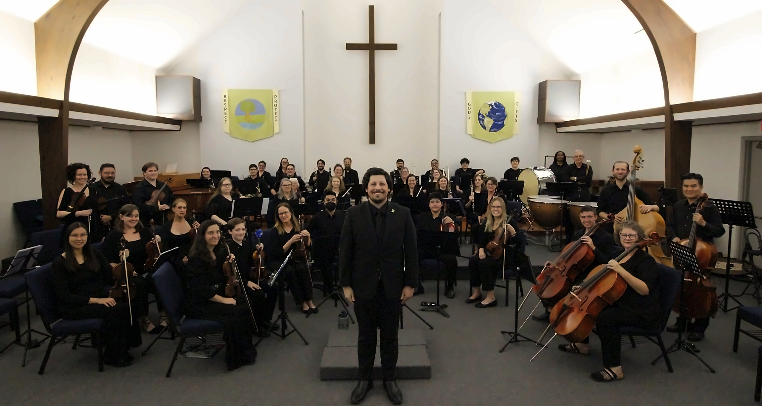 Symphony HEB orchestra seated in the sanctuary with the conductor standing at center, smiling at the camera